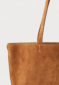 Tote-Bag aus tanfarbenem Wildleder mit glatter Textur, doppelten Ledergriffen und kontrastierender Naht entlang der Kanten. Schlichte, rechteckige Form.