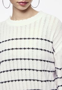 Biały dziany sweter z poziomymi czarnymi paskami. Wzór obejmuje dekolt z falbanką oraz fakturowany wzór na całej powierzchni.