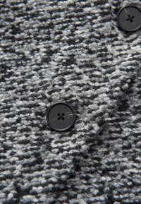 Tejido de tweed gris y negro con una superficie texturada, que presenta botones negros prominentes, redondos y con cuatro agujeros.