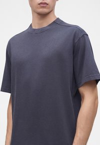 Jeune homme portant un t-shirt à manches courtes gris foncé uni avec un petit logo "BOSS" sur l'encolure avant.