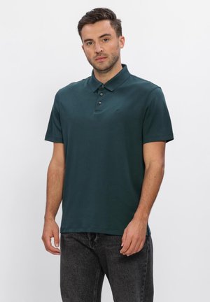 Man staat en draagt een donker groene polo met korte mouwen en een zwarte spijkerbroek tegen een effen lichte achtergrond.