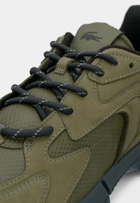 Olive green sneaker with a textured mesh upper and suede accents. Features black laces, a reinforced toe, and a logo near the tongue.
