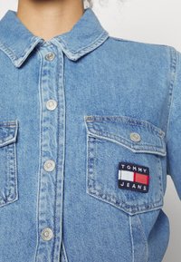 Světle modrá denimová košile s špičatým límcem, patentkami a dvěma předními kapsami. Obsahuje vyšitý logo nášivku "TOMMY JEANS" na levé kapse.