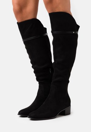 Bottes en suède noir hautes avec une texture lisse, dotées d'une fermeture zippée, d'un talon bloc solide et d'un accent en cuir décoratif.