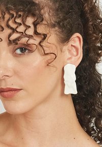 Baltas sveķu auskari ar teksturētu, abstraktu formu, veidoti karājošā dizainā, kas papildina nēsātāja iezīmes.