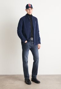 Camicia button-up in velluto a coste blu navy, dolcevita nera, jeans di denim scuro, stivali neri e un cappello blu con un design rosso e bianco.
