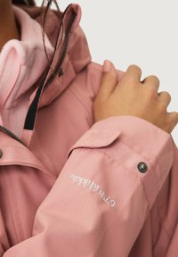 Chaqueta impermeable rosa con cuello alto, cierre de cremallera y un suave forro rosa. Presenta un puño con botón a presión y detalle del logotipo bordado.