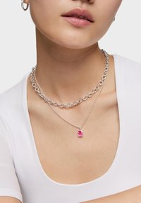 Donna che indossa due collane d'argento, una con una catena spessa e l'altra con un piccolo ciondolo a forma di orsacchiotto gommoso rosa, abbinata a una maglietta bianca.