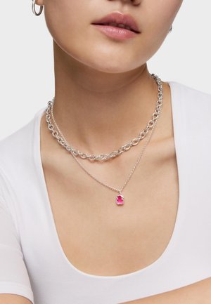 Kobieta nosząca dwa srebrne naszyjniki, jeden gruby łańcuch, a drugi z małą różową zawieszką w kształcie misia żelka, w połączeniu z białą bluzką.