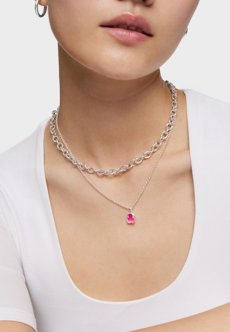 Donna che indossa due collane d'argento, una con una catena spessa e l'altra con un piccolo ciondolo a forma di orsacchiotto gommoso rosa, abbinata a una maglietta bianca.