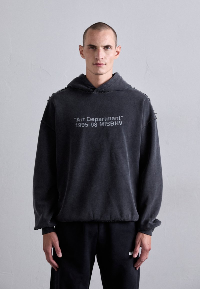 Mann mit kurzen Haaren, der einen verwaschenen schwarzen Hoodie mit dem Text "Art Department 1995-08 MISBHV" trägt, steht vor einem schlichten hellgrauen Hintergrund.
