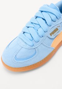 Light blue suede sneaker with round toe, flat laces, orange accents, and a textured rubber sole. Features logo in gold on the side.