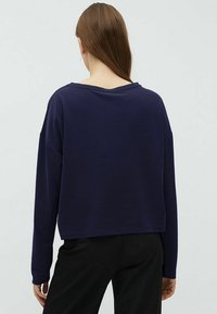 Námornícky modrý skrátený sveter s dlhými rukávmi a širokým výstrihom. Mäkká textúra, minimalistický dizajn. Nosený cez čierne nohavice.
