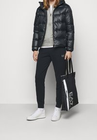 Doudoune noire, pull gris, pantalon de survêtement foncé, baskets blanches, sac fourre-tout noir avec logo EA7. Comprend une capuche et une fermeture éclair à l'avant.