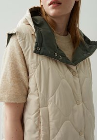 Beige gesteppte Weste mit grüner Kapuze. Verfügt über Druckknöpfe und seitliche Taschen. Weiche Textur und wellenförmiges Muster auf dem Stoff.