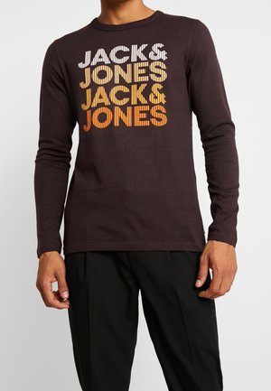 Mies, który ma na sobie ciemno brązową koszulę z długim rękawem z napisem "JACK & JONES" wydrukowanym trzy razy w białym, żółtym i pomarańczowym gradiencie kolorów.