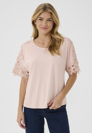 Pink T-shirt with intricate lace detailing on the sleeves and a round neckline, made from soft fabric. Paired with dark denim jeans.