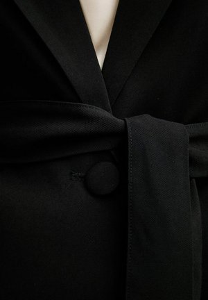 Cintura in tessuto nero annodata intorno a un cappotto nero con un bottone rotondo coperto e uno strato interno beige visibile all'altezza del collo.