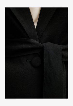 Cintura in tessuto nero annodata intorno a un cappotto nero con un bottone rotondo coperto e uno strato interno beige visibile all'altezza del collo.