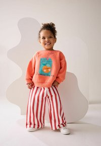 Koralfarvet sweatshirt med cartoon juice box grafik, parret med rød og hvid stribet bred benbukser og hvide sneakers. Glat stoftekstur.