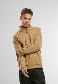 Hnedý pulóver na zips vyrobený z mäkkého materiálu, s náprsním vreckom na zips, bočnými vreckami a textúrovanými akcentmi na pleciach.