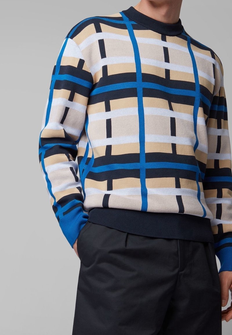 Homme portant un pull à manches longues à carreaux beige, bleu, noir et blanc avec un pantalon noir, main dans la poche, sur un fond gris uni.