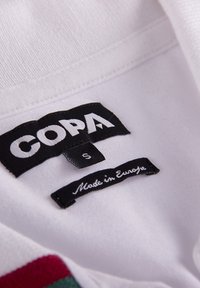 Tissu blanc avec une étiquette montrant "COPA" en lettres blanches grasses, la taille "S" imprimée en gris, et "Fabriqué en Europe" en écriture cursive sur une étiquette noire.