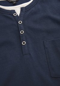 Mörkblå bomullshenley med vit krage, fyra grå knappar och en bröstficka. Texturen är slät och något elastisk.