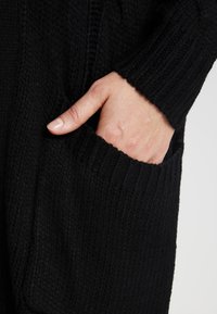Cardigan nero lavorato a maglia con texture a coste, maniche lunghe e una tasca laterale con una mano inserita.