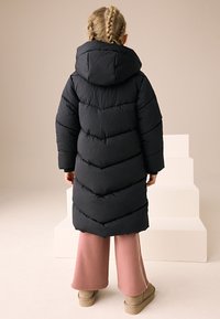 Manteau matelassé noir avec capuche, présentant une texture matelassée en chevron. Porté sur un pantalon large rose et des bottines beiges.