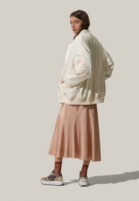 Giacca bomber in satin crema, gonna midi in satin color sabbia, sneakers marroni con motivo e dettagli bianchi, e calzini a coste color bordeaux visibili.