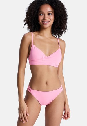 Femme aux cheveux bouclés souriant, portant un bikini rose avec des fines bretelles et des bandes doubles sur les côtés du bas, sur un fond uni.