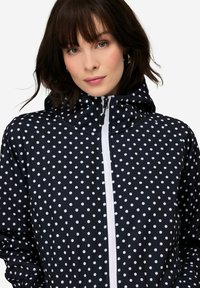 Mustapilkullinen takki hupulla, jossa on valkoinen etuvetoketju ja sileä, kiiltävä pinta. Kuosi koostuu tasaisesti sijoitetuista valkoisista palloista.