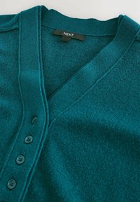 Teal cardigan lavet af blødt tekstureret stof, med V-hals, fem runde knapper og en syet kant design. Brandmærket er synligt.