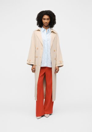 Femme aux cheveux bouclés portant un trench-coat beige, une chemise bleu clair à motifs, un pantalon large rouge et des talons pâles, debout devant un fond blanc.