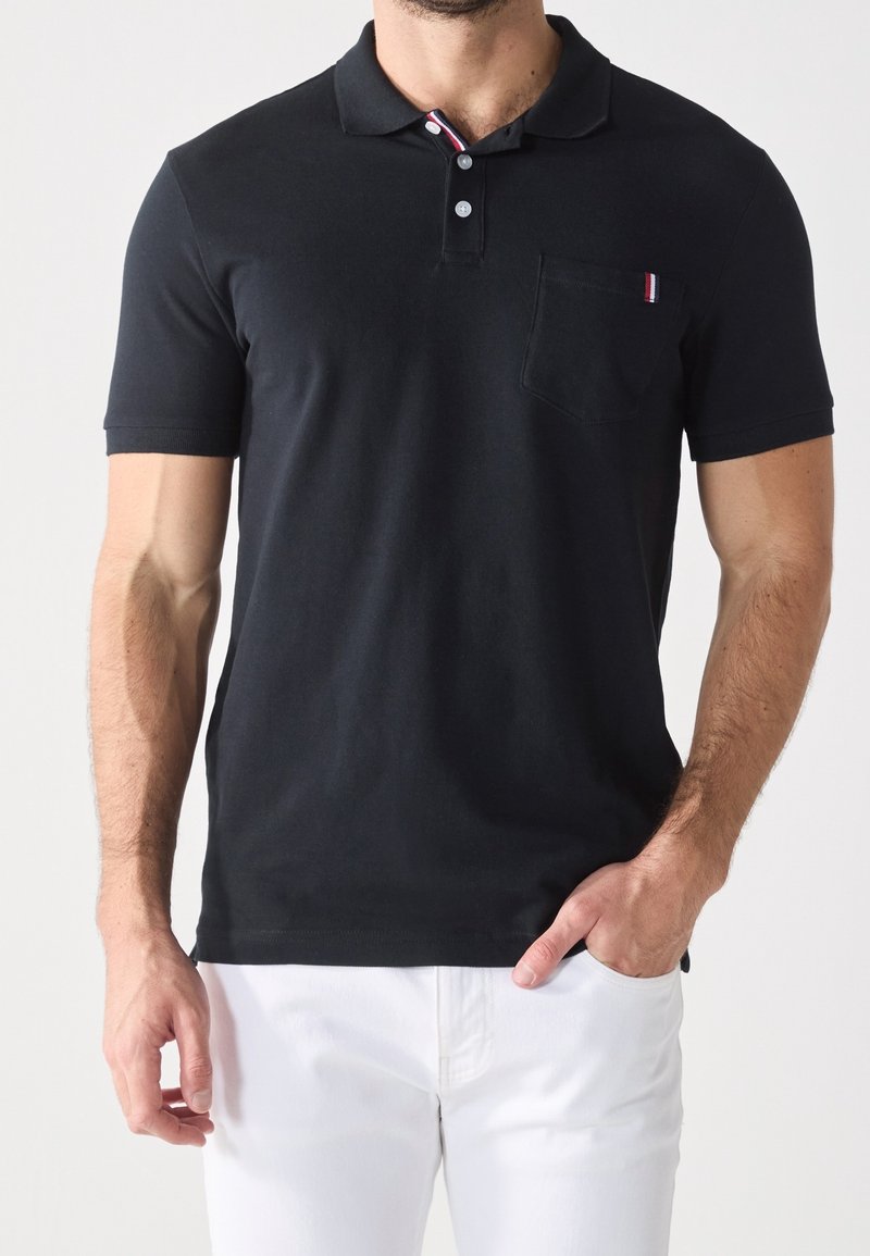 Homme portant un polo noir à manches courtes avec deux boutons et un petit détail de poche, associé à un pantalon blanc, main dans la poche.
