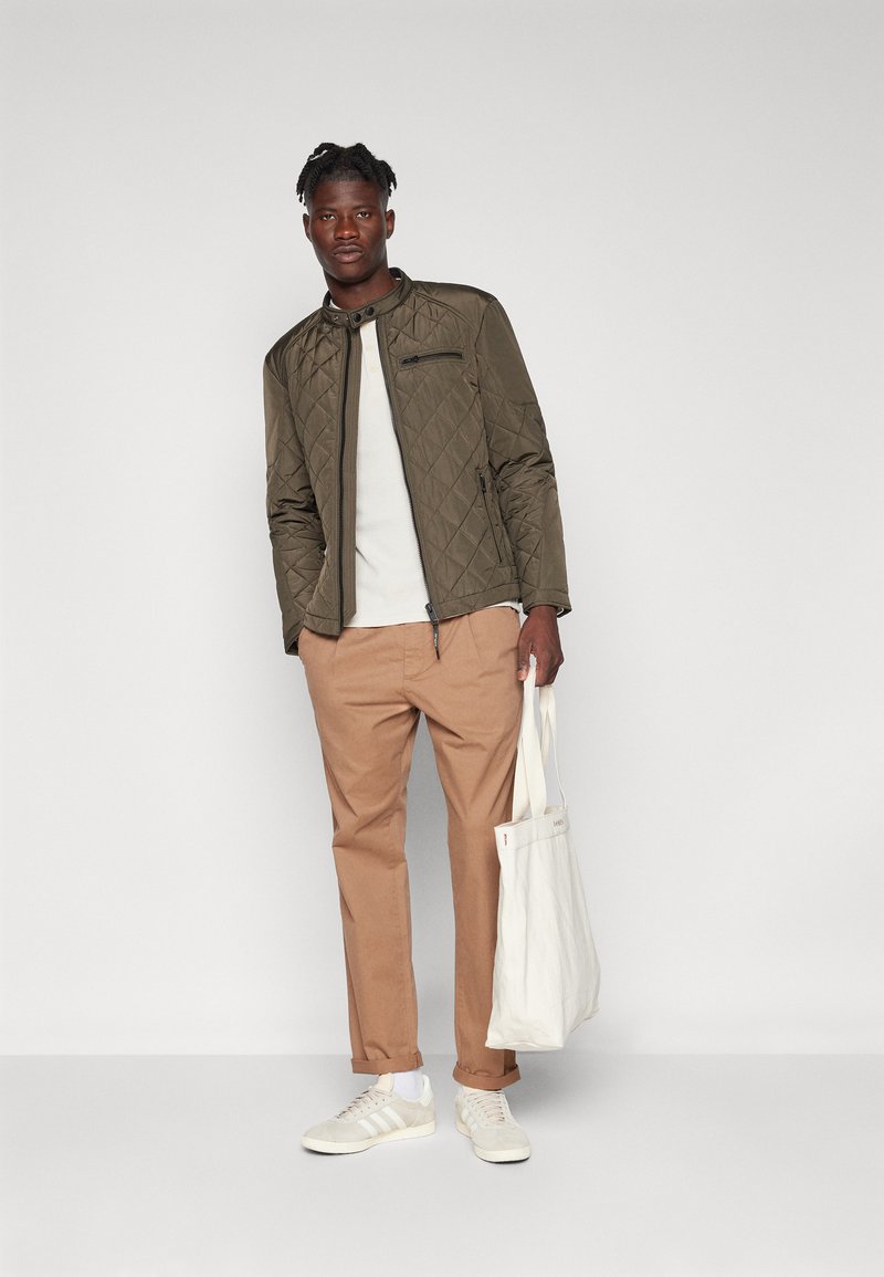 Gepolstertes olivgrünesJackett mit Stehkragen, beiger Pullover, hellbraune Hose und helle Sneaker. Trägt eine weiße Stofftasche.