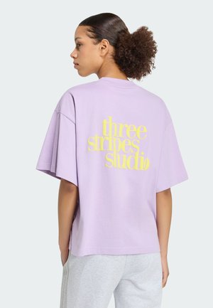 Ung kvinde med krøllet hår iført en lilla oversized t-shirt med gul "three stripes studio" tekst bagpå, iført lysegrå bukser.