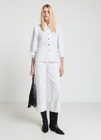 Weißes Hemd mit Puffärmeln, kombiniert mit weißen Hosen im weiten Bein. Schwarze Ledertasche mit Fransen und kniehohe schwarze Cowboy-Stiefel.