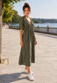 Donna con i capelli raccolti in uno chignon che indossa un vestito midi verde oliva e scarpe da ginnastica bianche, in piedi su una passeggiata soleggiata lungo il fiume con alberi e ringhiera.
