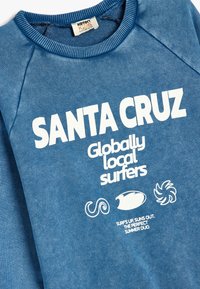 Niebieska bluza z białym napisem "SANTA CRUZ Globally Local Surfers". Posiada okrągły dekolt i rękawy raglanowe, miękka tekstura, casualowy design.