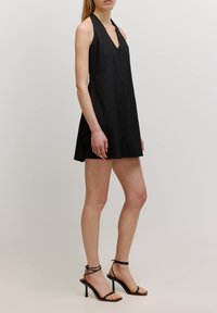Schwarzes Halterkleid mit V-Ausschnitt, A-Linien-Silhouette und knielangem Saum. Getragen mit schwarzen, schmalen, hochhackigen Sandalen. Glattes Material.
