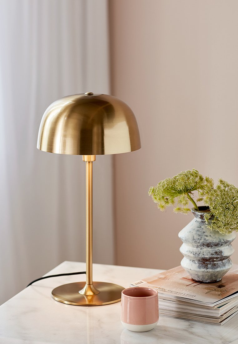 Messingtischlampe mit einem runden Schirm, einem schlanken Gestell und einem kreisförmigen Fuß. In der Nähe befindet sich eine rosa Keramiktasse und eine strukturierte Vase mit Grünpflanzen.