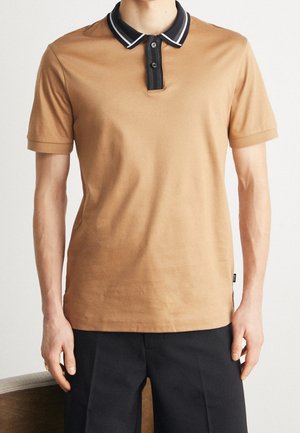 Mężczyzna w beżowej koszulce polo z krótkim rękawem, czarnym kołnierzykiem i listwą z guzikami, ubrany w czarne spodnie, stoi na jednolitym tle.