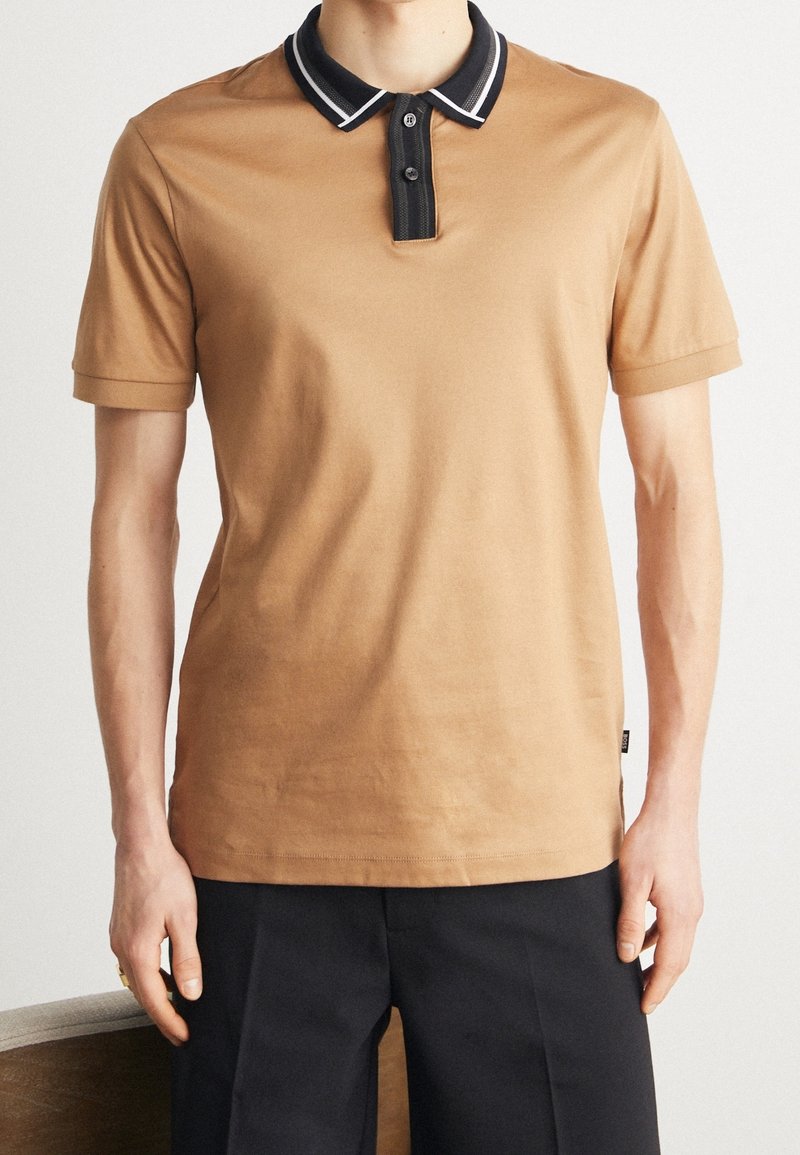 Homme portant un polo à manches courtes beige avec un col et une patte de boutonnage noirs, associé à un pantalon noir, debout devant un fond uni.