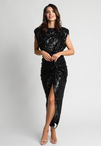 Kobieta z brązowymi włosami, ubrana w czarną, bezrękawnikową sukienkę z cekinami z rozcięciem z przodu oraz beżowe sandały na wysokim obcasie, stojąca na jednolitym tle.