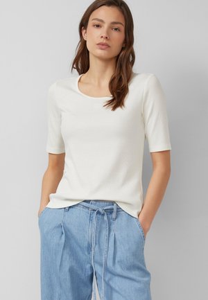 Femme aux cheveux bruns portant un haut blanc à manches courtes et un jean bleu clair taille haute avec une ceinture nouée, mains dans les poches, fond neutre.