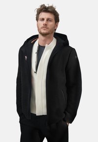 Uomo con giacca con cappuccio nera sopra un maglione bianco con zip e pantaloni scuri, in piedi con le mani nelle tasche su uno sfondo chiaro.