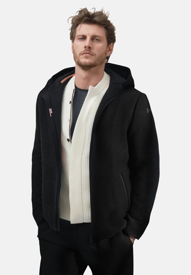 Uomo con giacca con cappuccio nera sopra un maglione bianco con zip e pantaloni scuri, in piedi con le mani nelle tasche su uno sfondo chiaro.