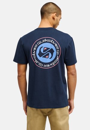 Mężczyzna zwrócony tyłem, noszący granatowy t-shirt z dużym okrągłym logo Quiksilver w kolorach niebieskim, różowym i czarnym na plecach, w połączeniu z beżowymi spodniami.
